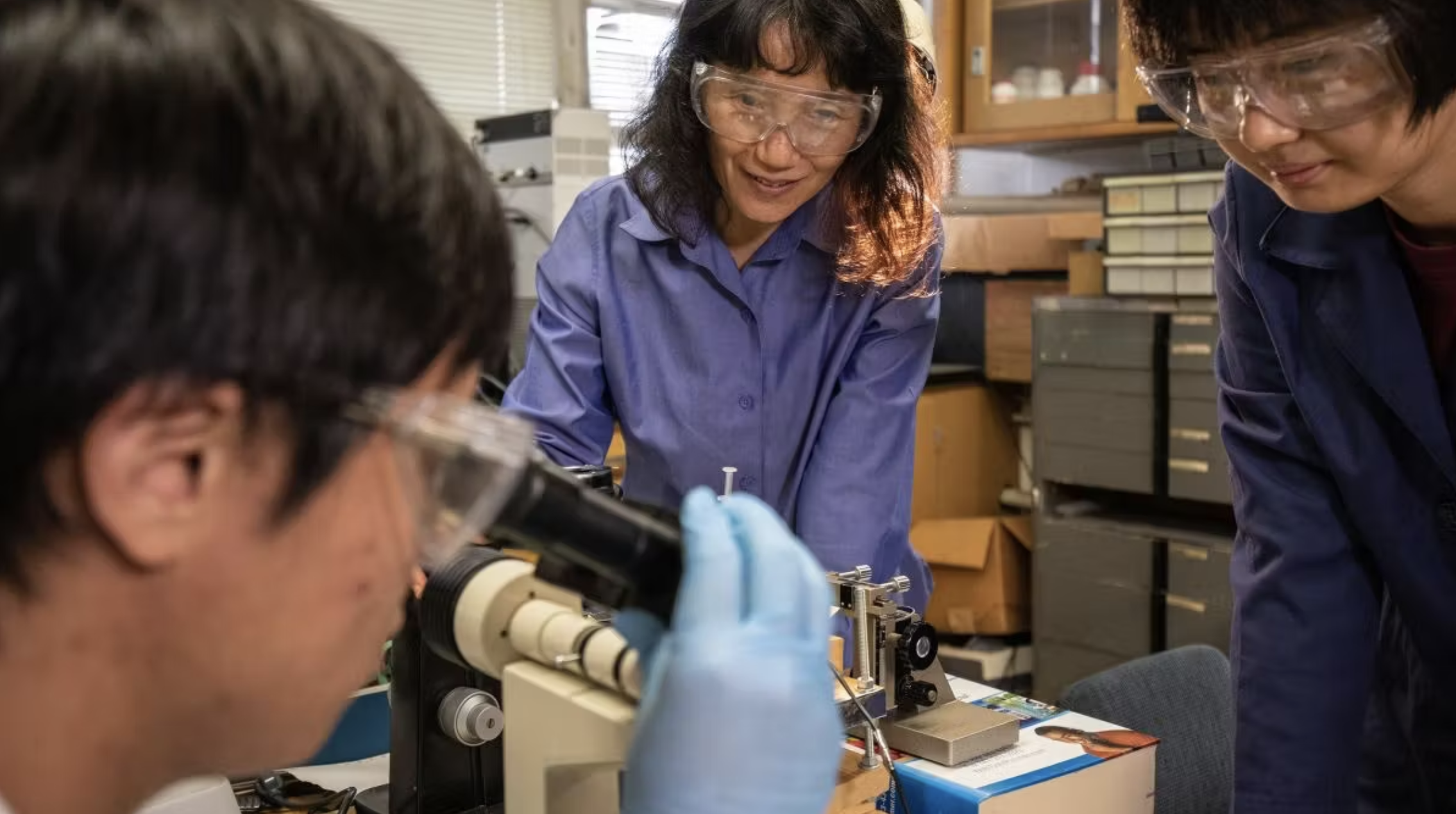 Hong Liang wears safety goggles and observes the students' work with a microscope.
